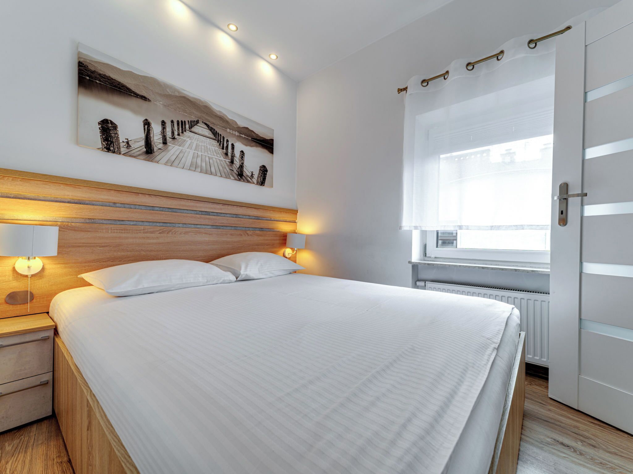 A modern hotel room with a wooden headboard, a large bed, bedside lamps, a window with curtains, and a wall-mounted artwork.