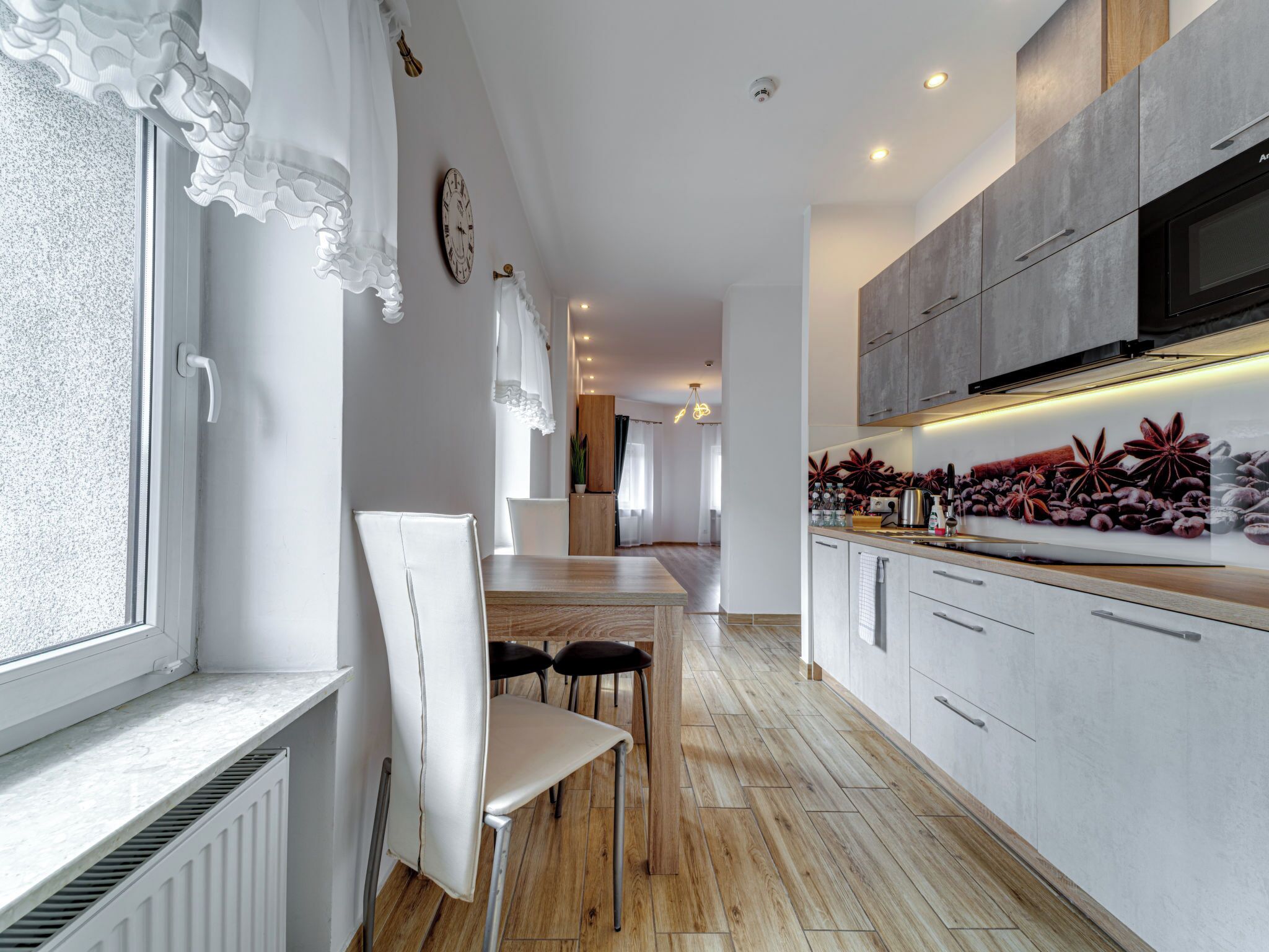 A modern kitchen with white cabinetry, a dining area with a wooden table and chairs, and a large window with white curtains.