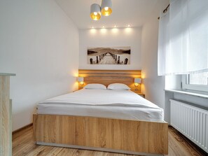 Un dormitorio moderno con una cama de madera, luces fijadas a la pared, un cuadro sobre la cama y una ventana con cortinas.