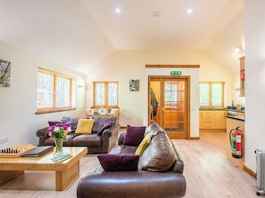 A spacious living area with a sofa, a coffee table, and a chessboard. There is a dining area with a table and chairs, and a kitchen visible through an open door. - Pine Marten Lodge by Interhome (Ballater)