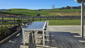 A wooden deck with a table and chairs, a white fence, and a view of green hills.
