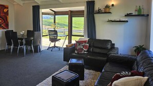 A living room with a black leather sofa, a dining table with chairs, a small coffee table, and a view of a green landscape through large windows.