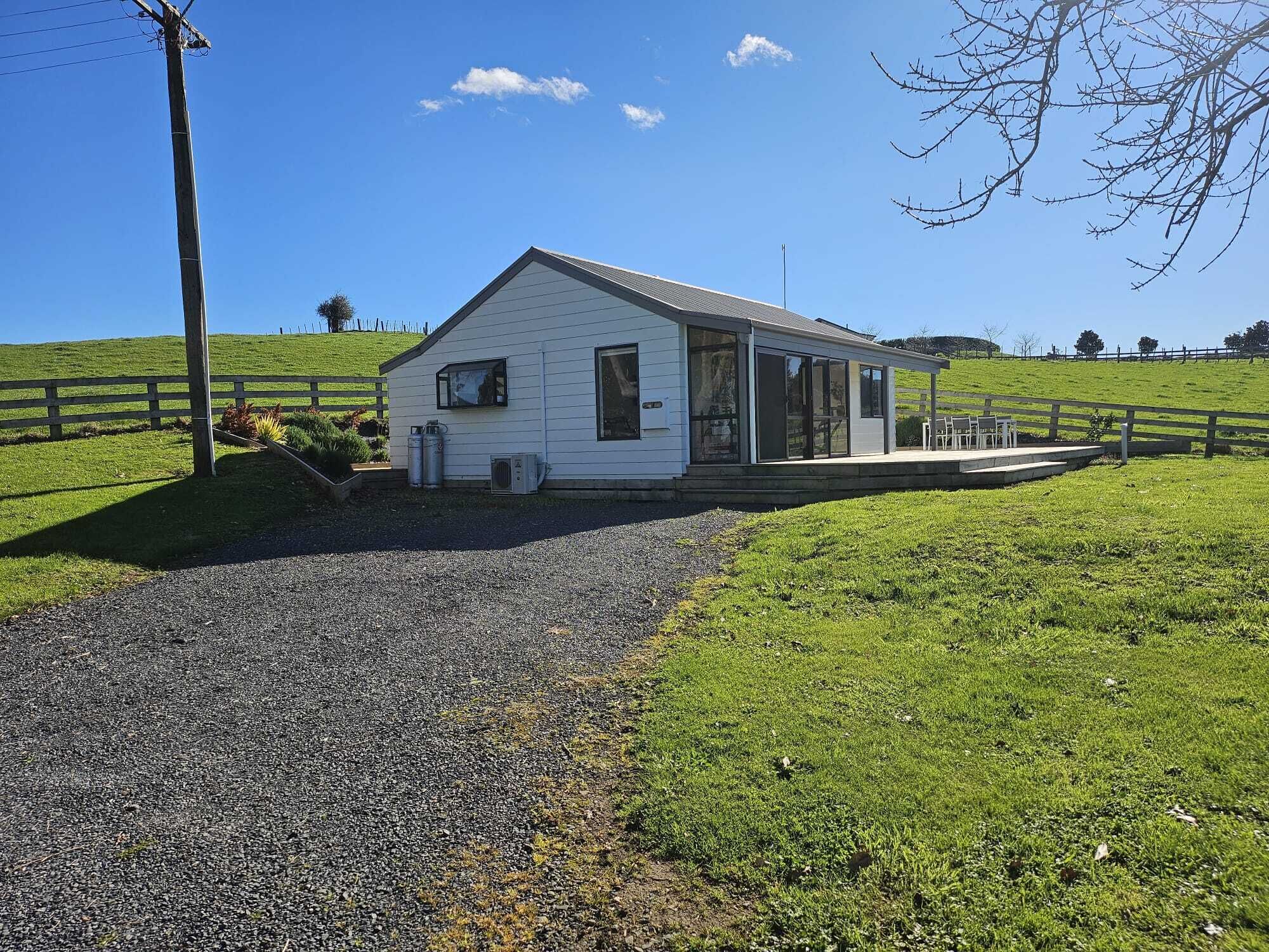 A white single-story building with a covered porch, a gravel driveway, and a grassy hill in the background.