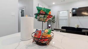 En korg med snacks står på en bänkskiva av marmor i ett modernt kök med en vit vägg och en platt-tv som är monterad ovanför bänkskivan.