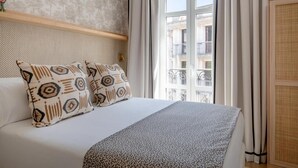Une chambre d’hôtel avec un lit, une fenêtre avec des rideaux, un objet décoratif rouge fixé au mur, et une vue sur un bâtiment à l’extérieur.