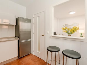 A kitchen with white tiles, a stainless steel refrigerator, two stools, and a mirror with a vase of yellow flowers. - Hommyhome San Esteban 001 (Sevilla)