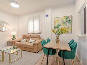 A modern living room with a beige sofa, a glass coffee table, and a dining table with green chairs.