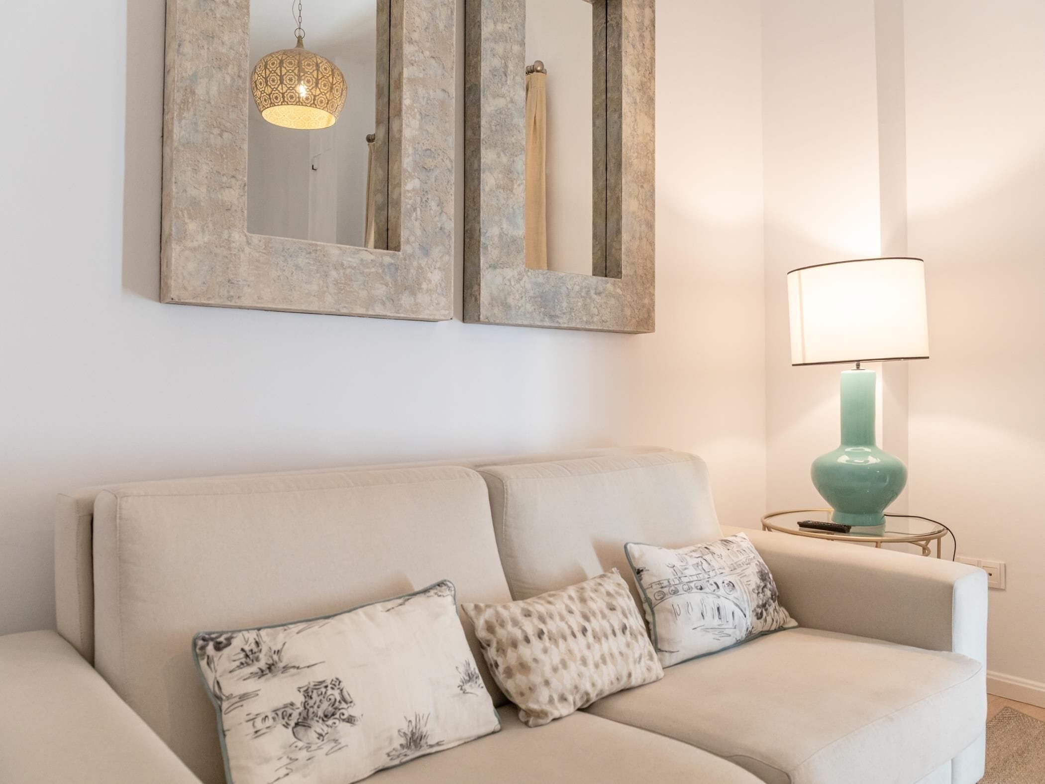 A neutral-toned living room with a sofa, patterned cushions, a hanging light fixture, and a wall-mounted mirror.