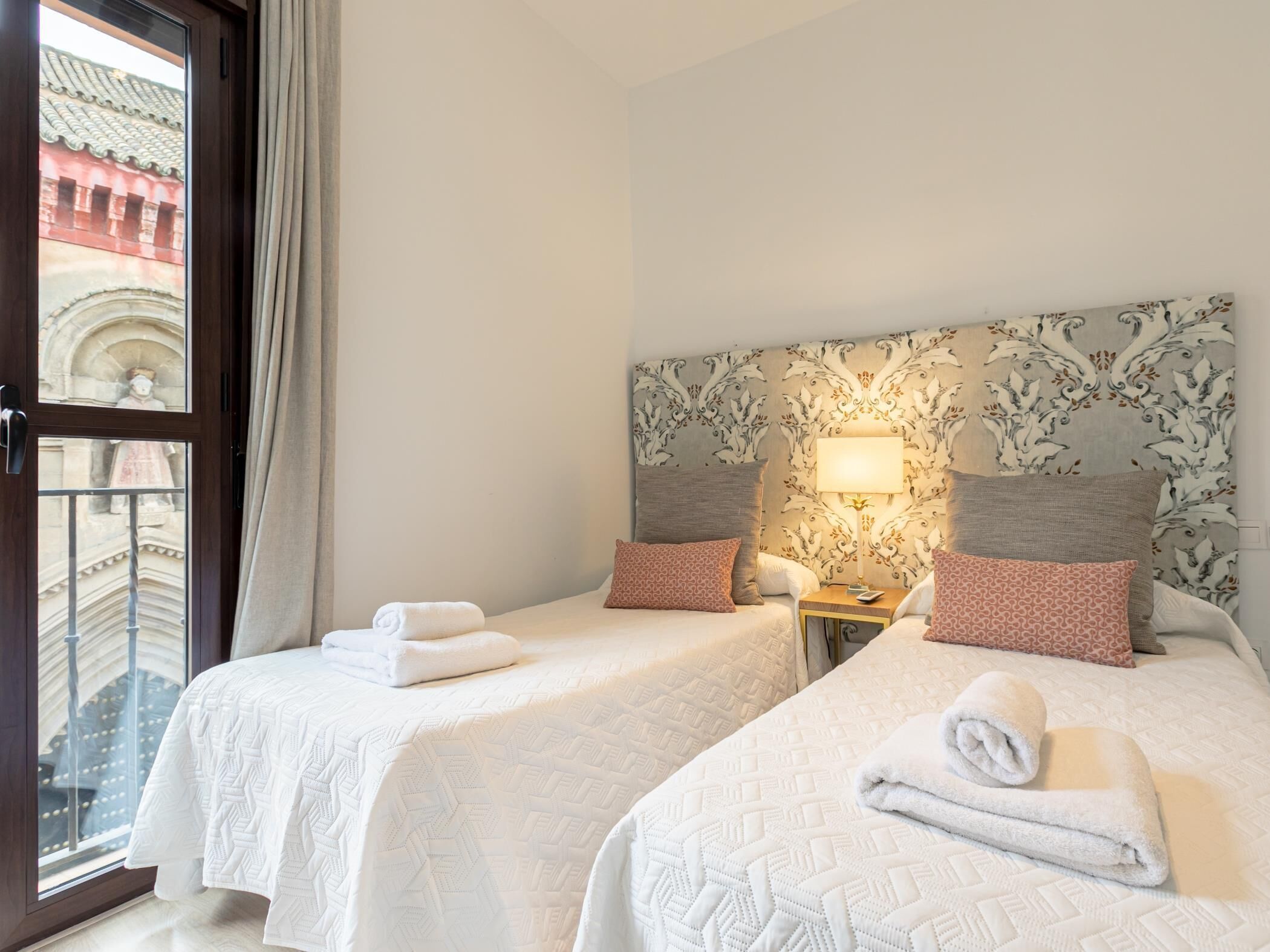 Een hotelkamer met twee bedden, een nachtkastje met een lamp en uitzicht op een gebouw door een glazen deur.