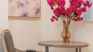 Une table ronde en bois, ornée d’un vase de fleurs roses, une chaise et un tableau accroché au mur.