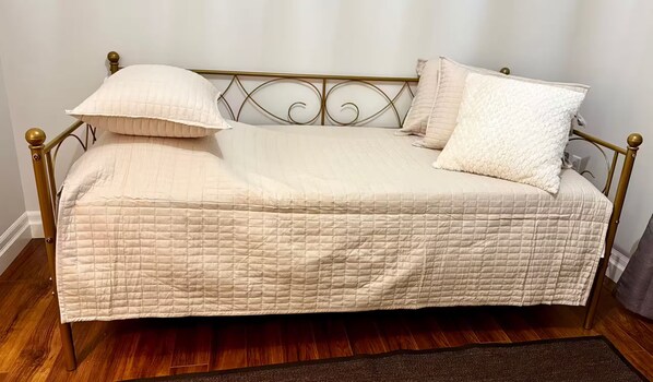 Un lit simple avec un matelas matelassé et deux oreillers, une tête de lit en métal et un plancher en bois foncé.