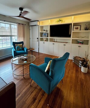 A living room with a blue armchair, a glass coffee table, a television on a white cabinet, and a ceiling fan.