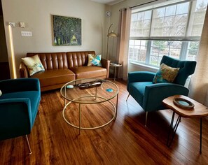 Un salon avec un canapé en cuir marron, des fauteuils turquoise, une table basse en verre et une grande fenêtre donnant sur un beau paysage.