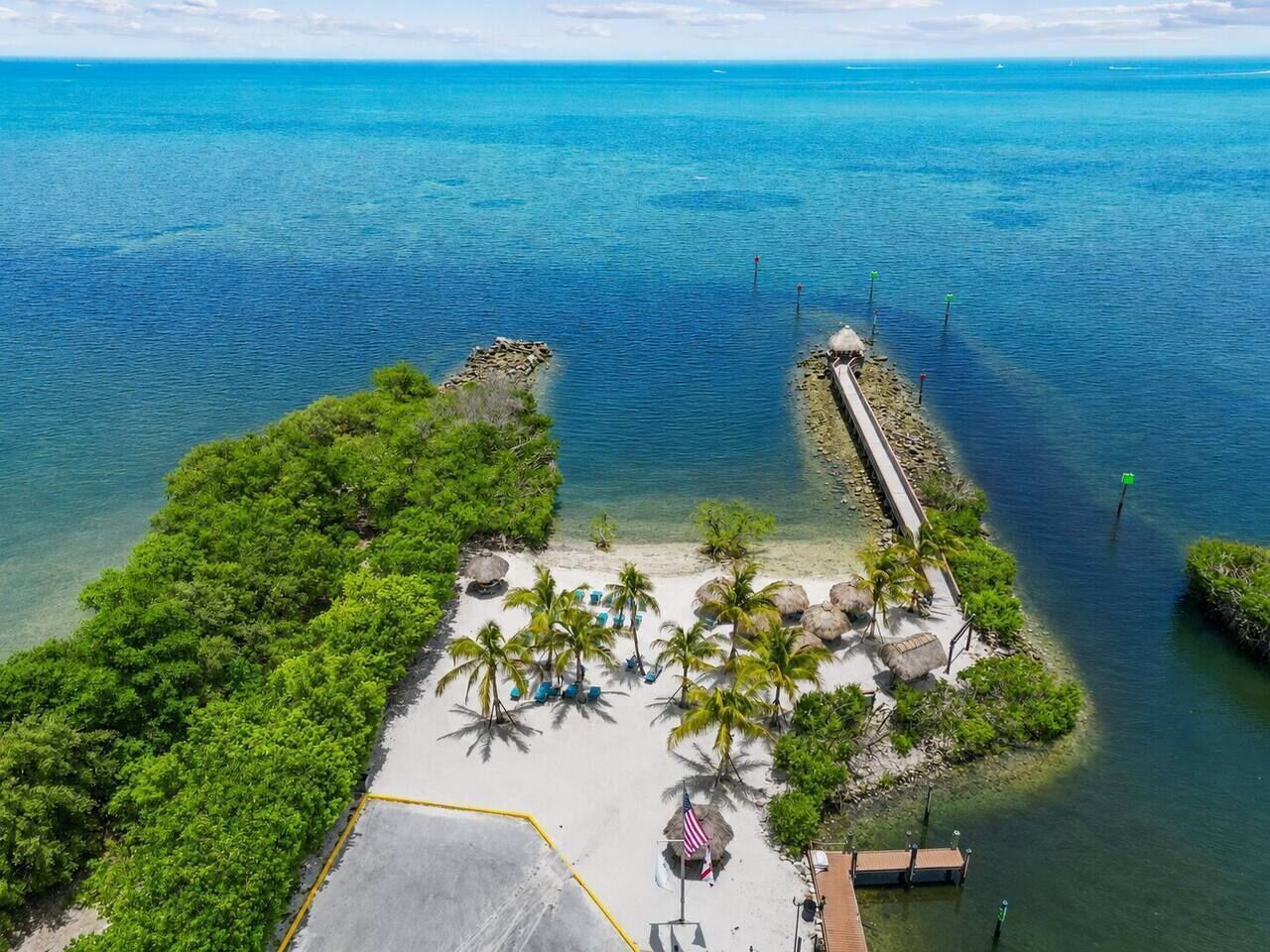 Vue aérienne d’un complexe hôtelier tropical situé sur une plage, avec une promenade dallée, des palmiers et une jetée qui s’avance dans l’eau.