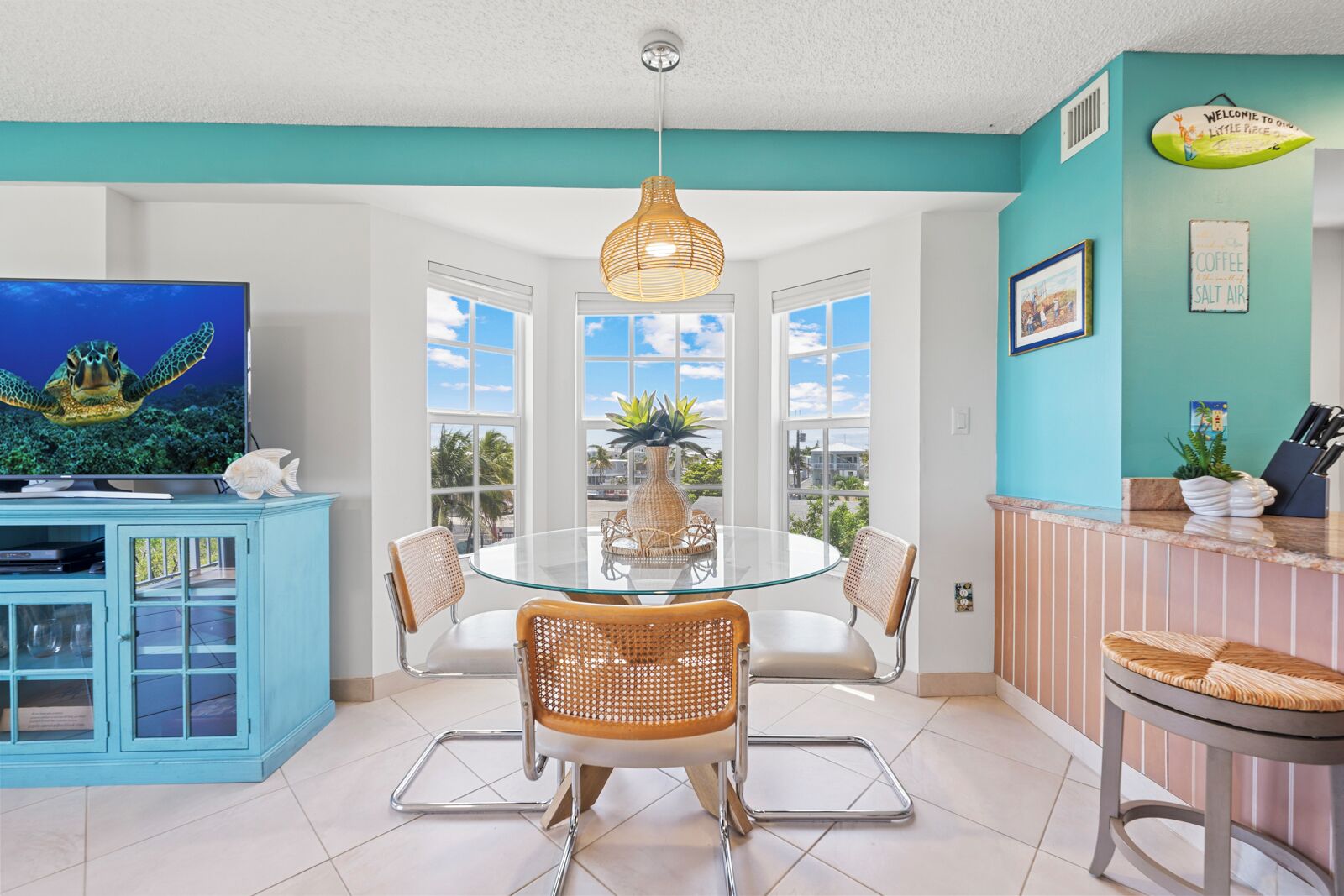 A modern kitchen with a round glass table, wicker chairs, and a blue cabinet with a TV.