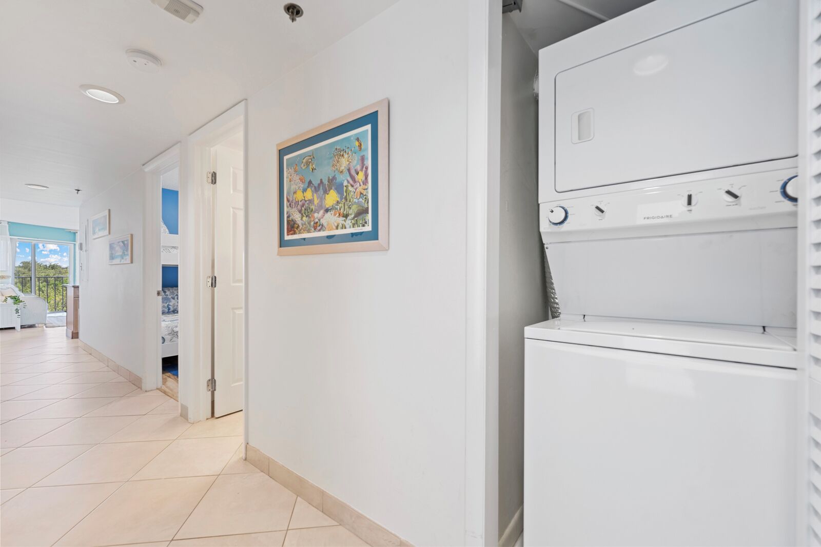 Un couloir d’hôtel avec une porte donnant sur une chambre, un tableau encadré accroché au mur et un appareil électroménager blanc, peut-être un lave-linge, dans un coin.