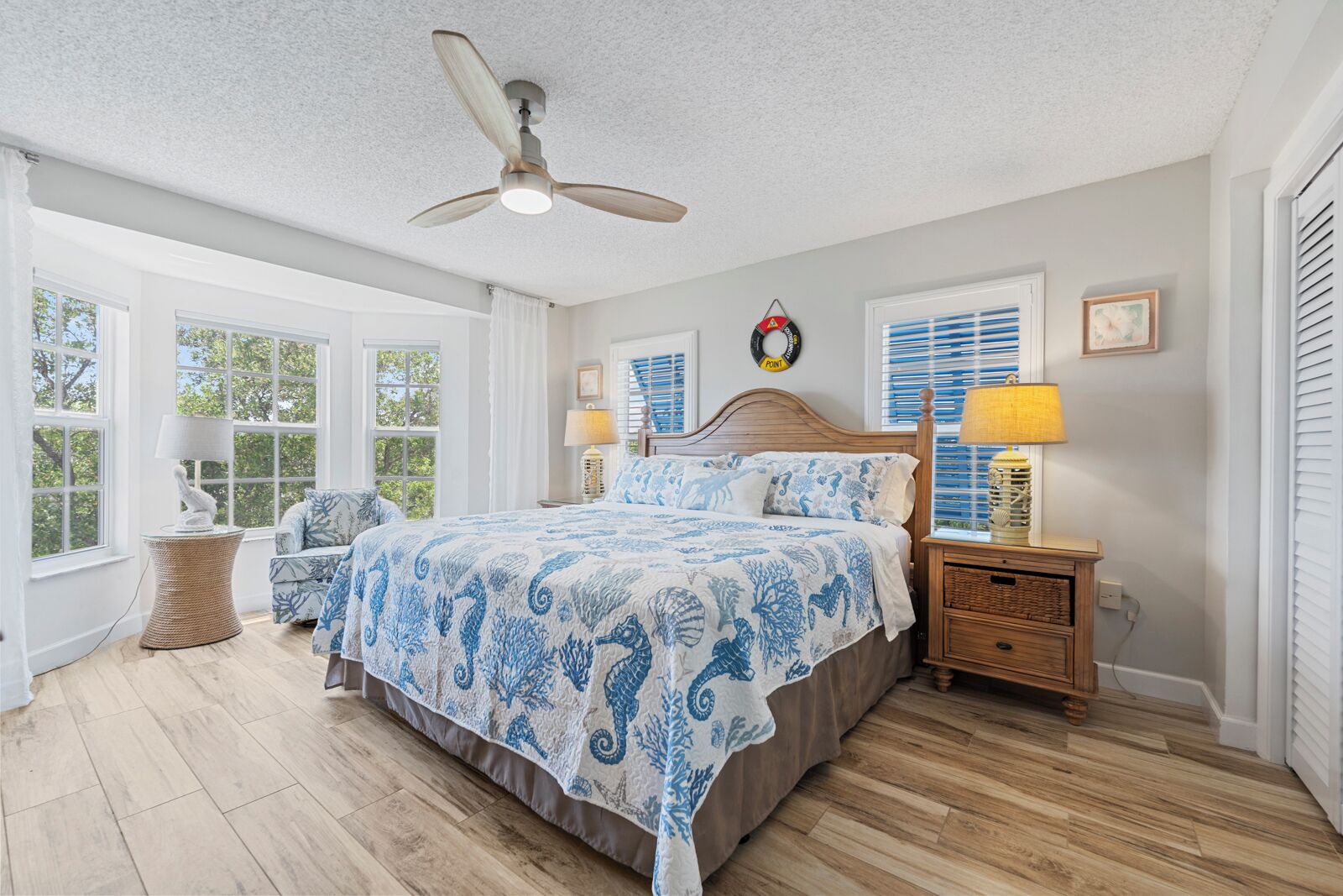 Une chambre à coucher comprenant un lit, une table de chevet, une lampe, un ventilateur de plafond et de grandes fenêtres.