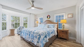 Une chambre à coucher comprenant un lit, une table de chevet, une lampe, un ventilateur de plafond et de grandes fenêtres.