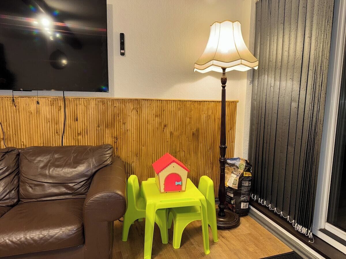 A room with a brown leather sofa, a small green table with a red house model, a lamp, and a window with blinds.
