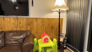 A room with a brown leather sofa, a small green table with a red house model, a lamp, and a window with blinds.