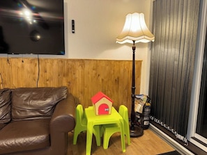 Una habitación con un sofá de cuero marrón, una mesita pequeña con un modelo de casa roja, una lámpara y una ventana con persianas.