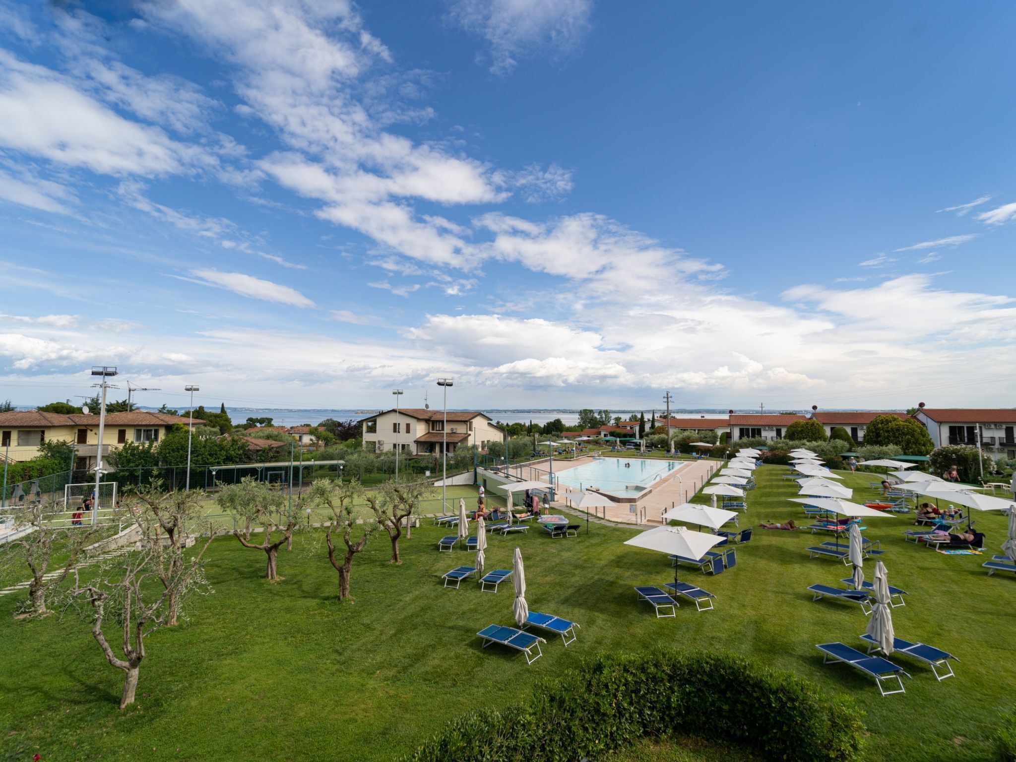A pool area with sun loungers and umbrellas, surrounded by greenery and buildings.