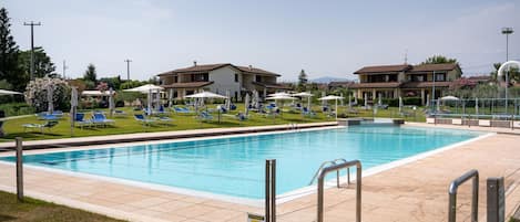 Une piscine entourée de bâtiments, avec des chaises longues et des parasols, sous un ciel dégagé.