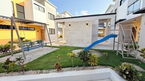 A modern residential area with a children's play area, including a swing set and a slide, surrounded by well-maintained grass and small plants. - Modern 2BR Oasis/Semi-Private Pool (Punta Cana)