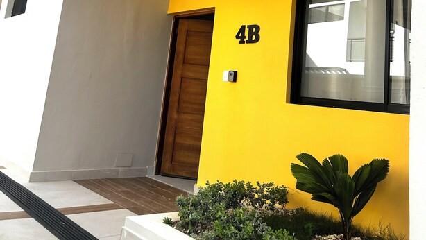 A modern building with a yellow facade, a wooden door, and a small garden area with plants. - Modern 2BR Oasis/Semi-Private Pool (Punta Cana)