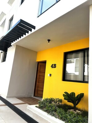A modern building with a yellow facade, a wooden door, and a small garden area with plants. - Modern 2BR Oasis/Semi-Private Pool (Punta Cana)