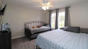 Ein Schlafzimmer mit einem Bett, einem zusätzlichen Bett, einem Deckenventilator, einem Fenster mit Vorhängen und einem Schreibtisch mit Ventilator.