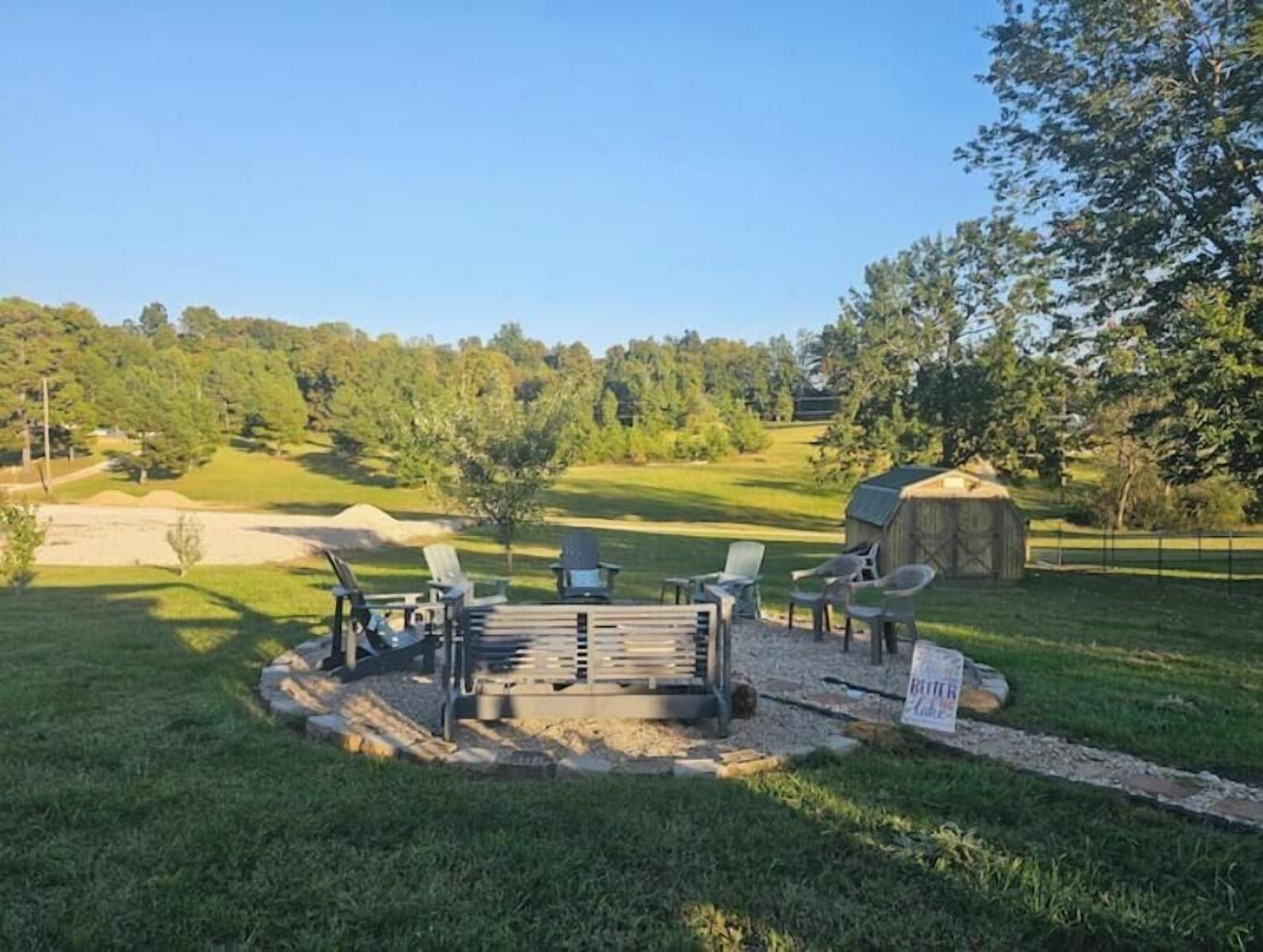 A circular outdoor seating area with chairs and a fire pit, surrounded by a grassy field and trees.