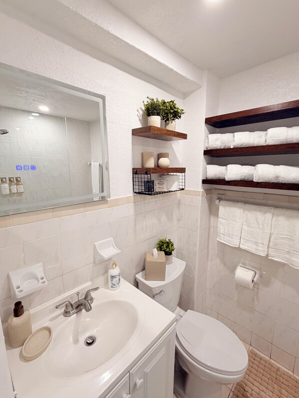 A bathroom with a white sink, mirror, toilet, and shelves with towels and toiletries.