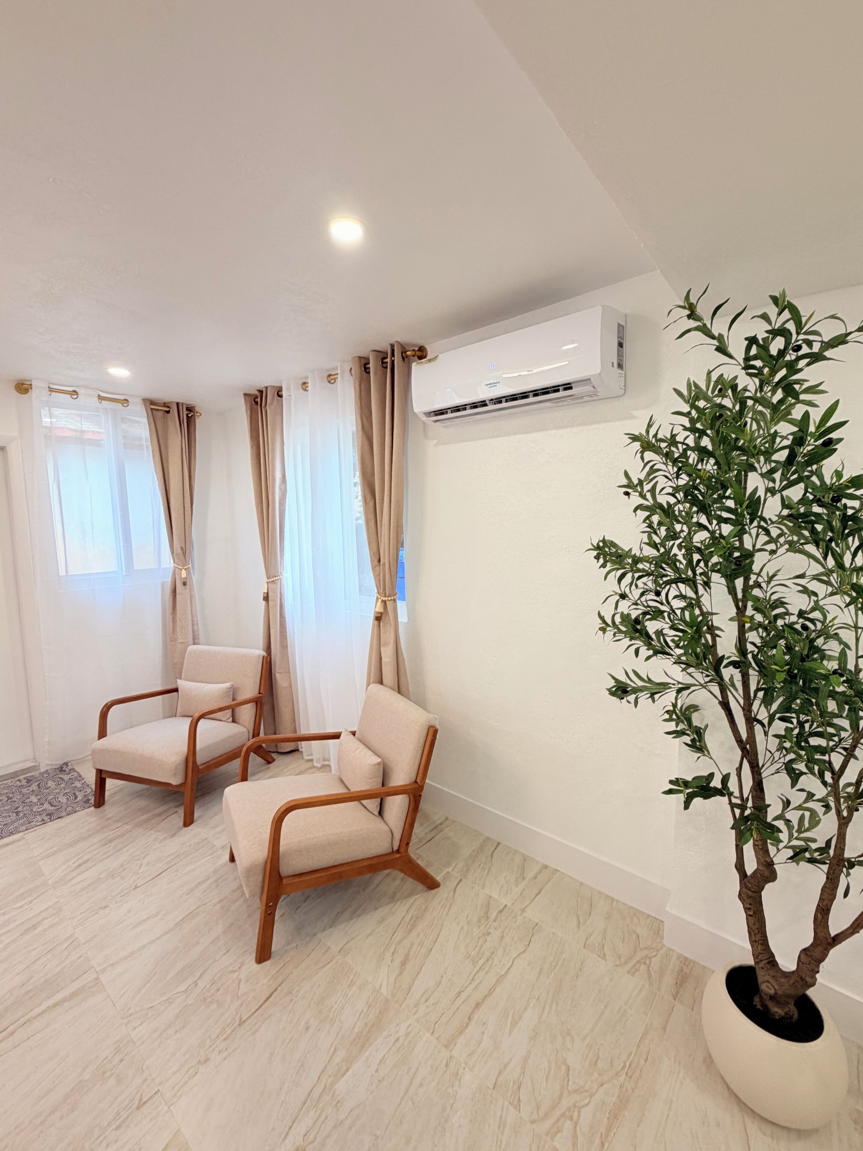 A room with two beige armchairs, a potted plant, and an air conditioning unit on the wall.