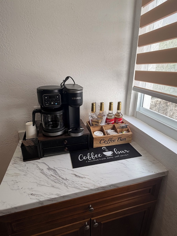 Un coin café aménagé avec une machine Keurig, des tasses à café et des sirops Torani, le tout posé sur un comptoir en marbre.