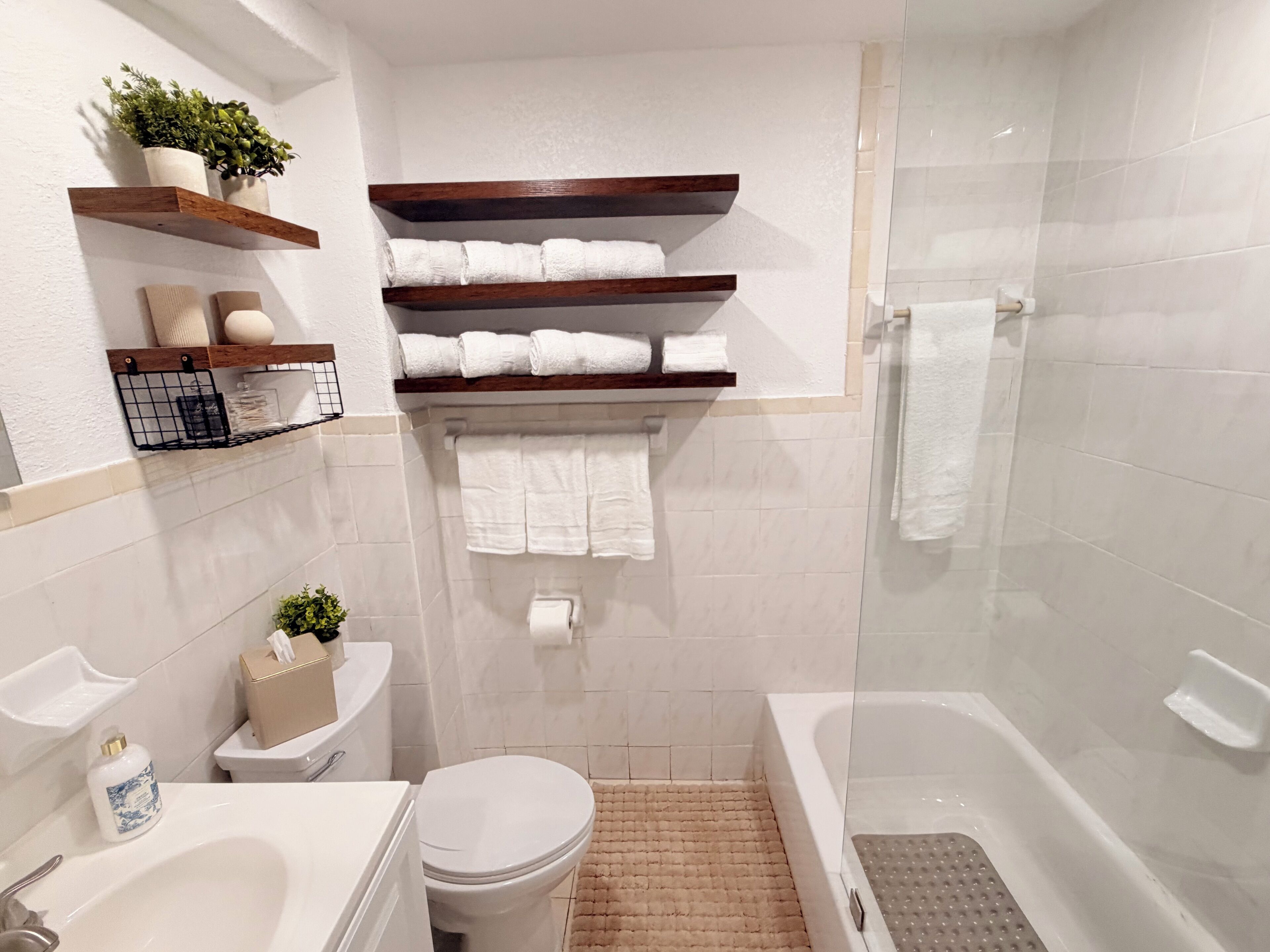 A modern bathroom with a toilet, sink, and bathtub. There are shelves with towels and a small plant.