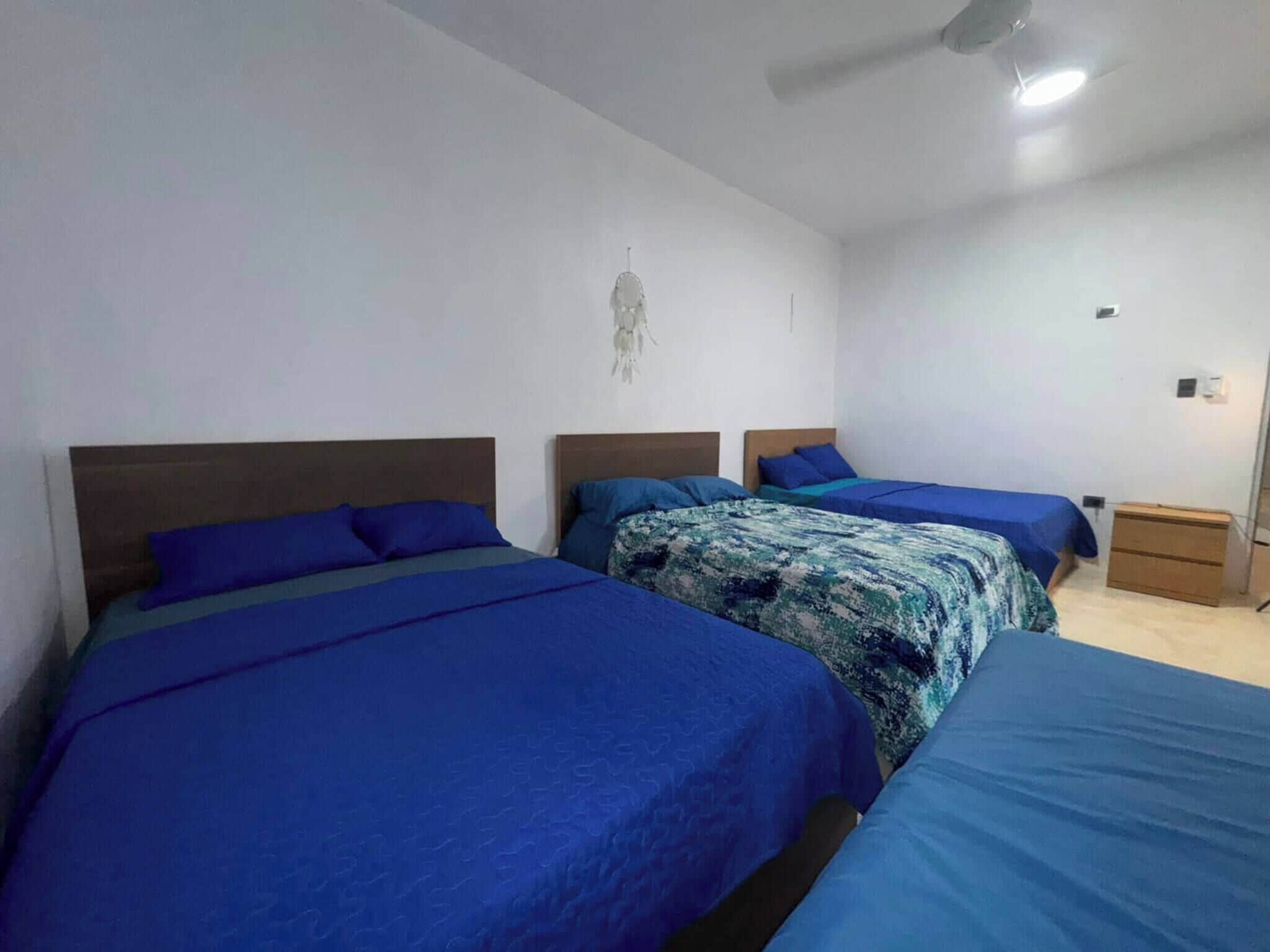 Una habitación con tres camas, cada una con ropa de cama azul, un ventilador de techo y un atrapasueños colgado en la pared.