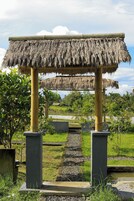 Estructura de paja con un sendero que lleva a un cuerpo de agua, rodeada de vegetación y un cielo despejado.