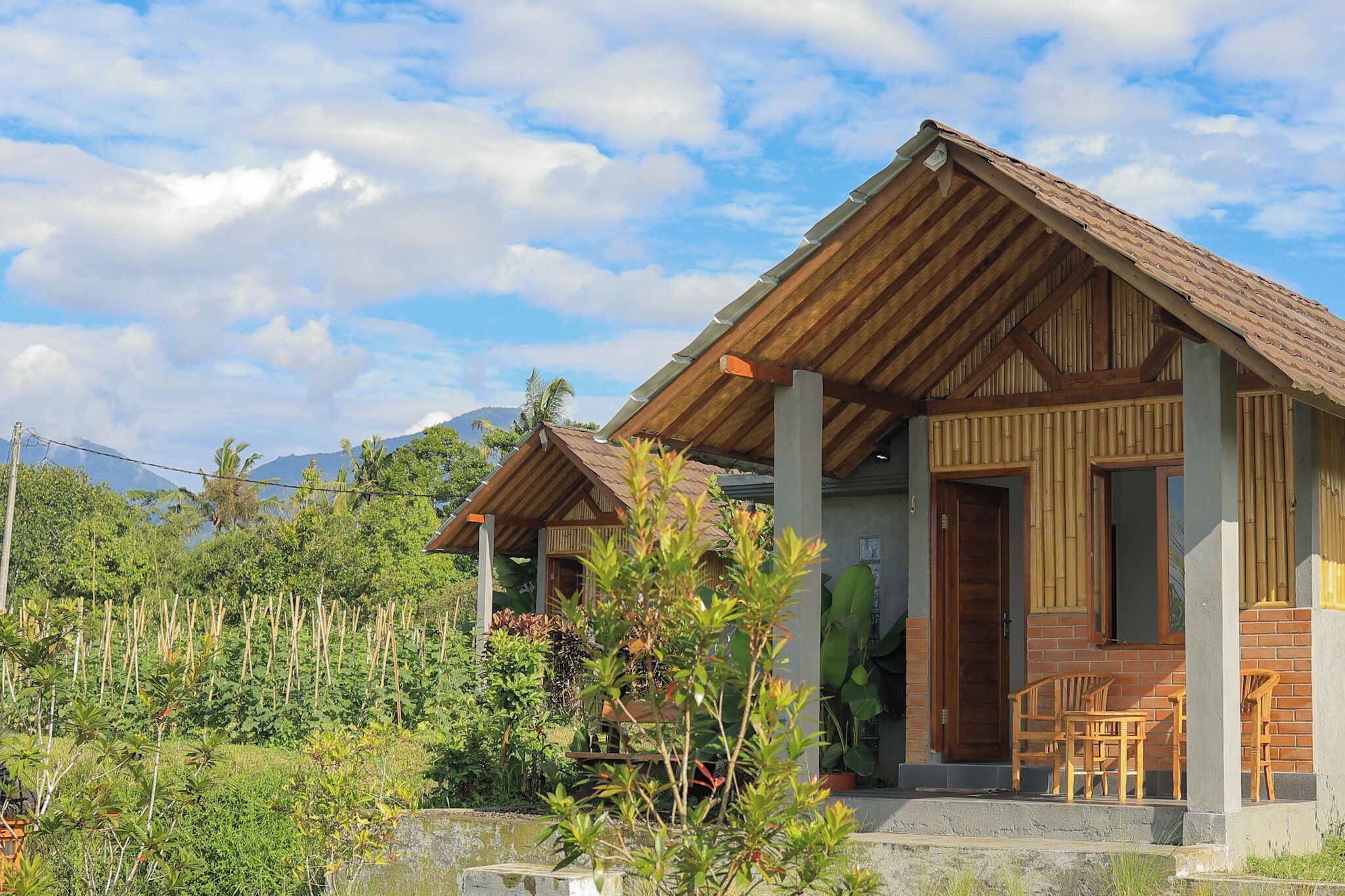 A bungalow with a wooden roof, a porch with chairs, and a view of a lush garden and mountains in the background.
