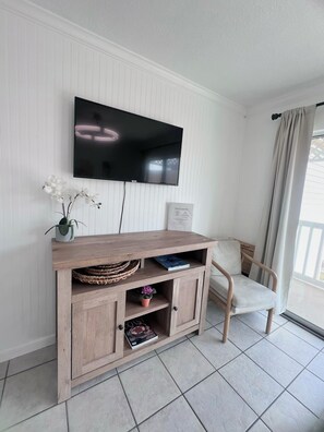 A room with a mounted flat-screen TV, a wooden entertainment unit with shelves, a potted plant, and a chair. - Coastal Oasis 1BR/1BA condo - Just steps from the Beach (Galveston)