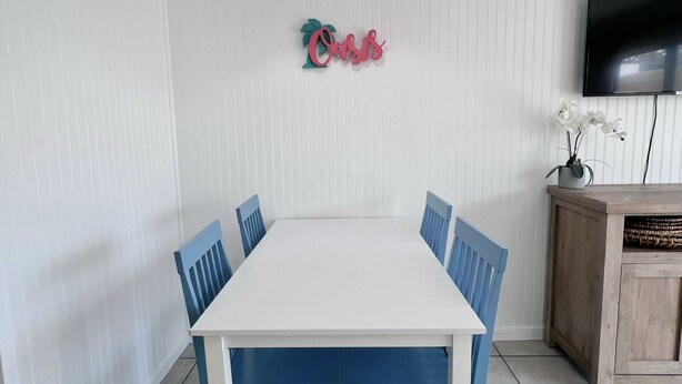 Zona pranzo con tavolo bianco e sedie blu, lampadario, televisore a schermo piatto e elemento decorativo a parete.