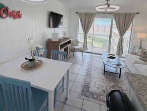 A modern living room with a dining area, a flat-screen TV mounted on the wall, and a balcony with curtains. - Coastal Oasis 1BR/1BA condo - Just steps from the Beach (Galveston)