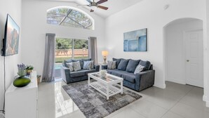 A spacious living room with a large window, a ceiling fan, a flat-screen TV, and a modern couch. - Beautiful 4bdr With Private Pool Gated Community (Almont)