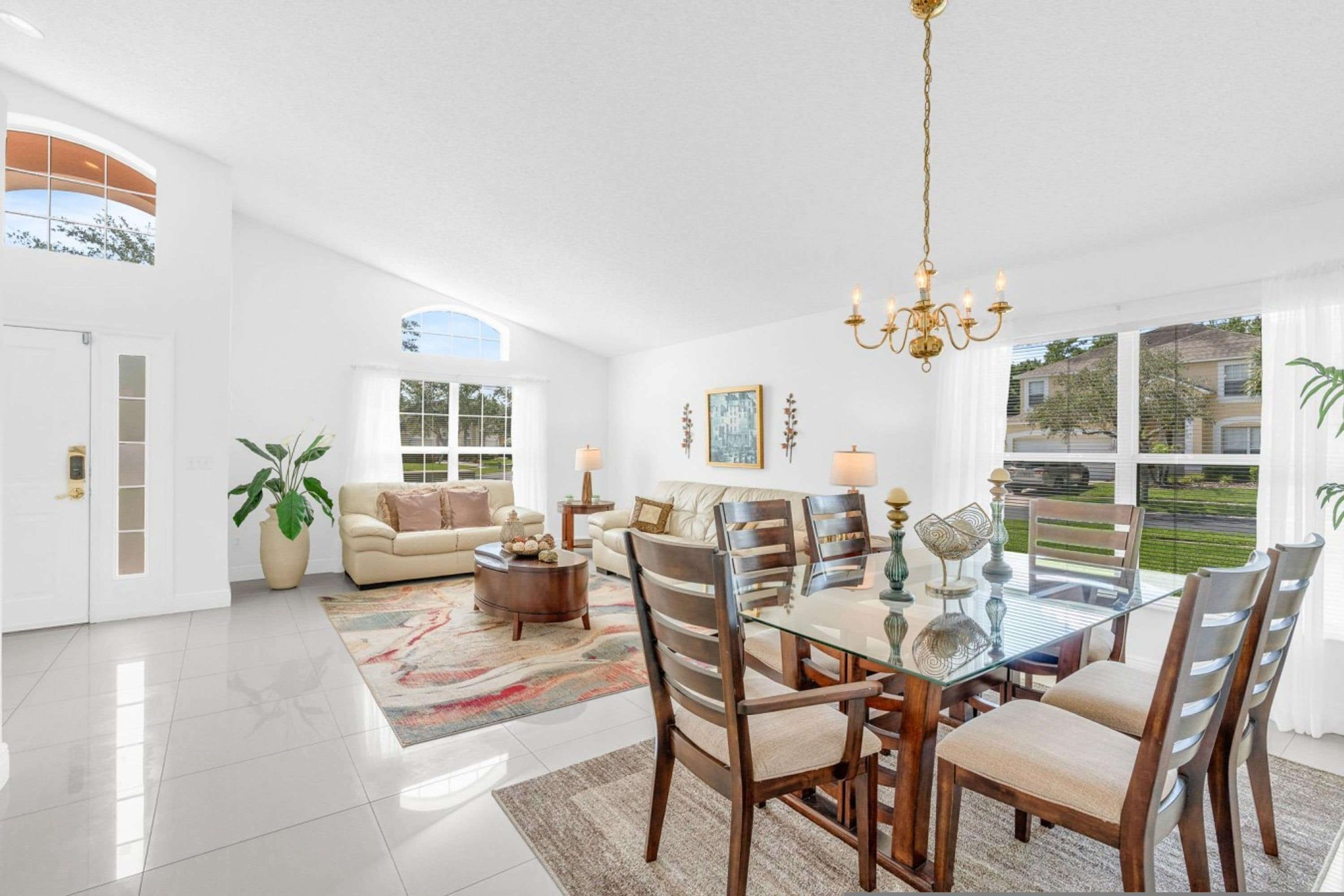 A spacious living room with a glass dining table, wooden chairs, a sofa, and a large window offering a view of the outdoors.