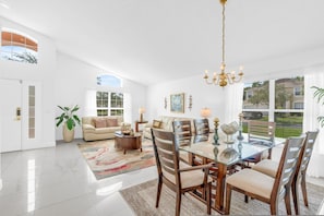 A spacious living room with a glass dining table, wooden chairs, a sofa, and a large window offering a view of the outdoors. - Beautiful 4bdr With Private Pool Gated Community (Almont)