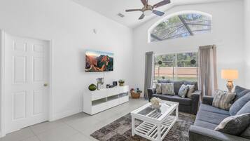 A spacious living room with a white ceiling fan, a large window with a view of the outdoors, a grey sofa, a white coffee table, and a flat-screen TV mounted on the wall.