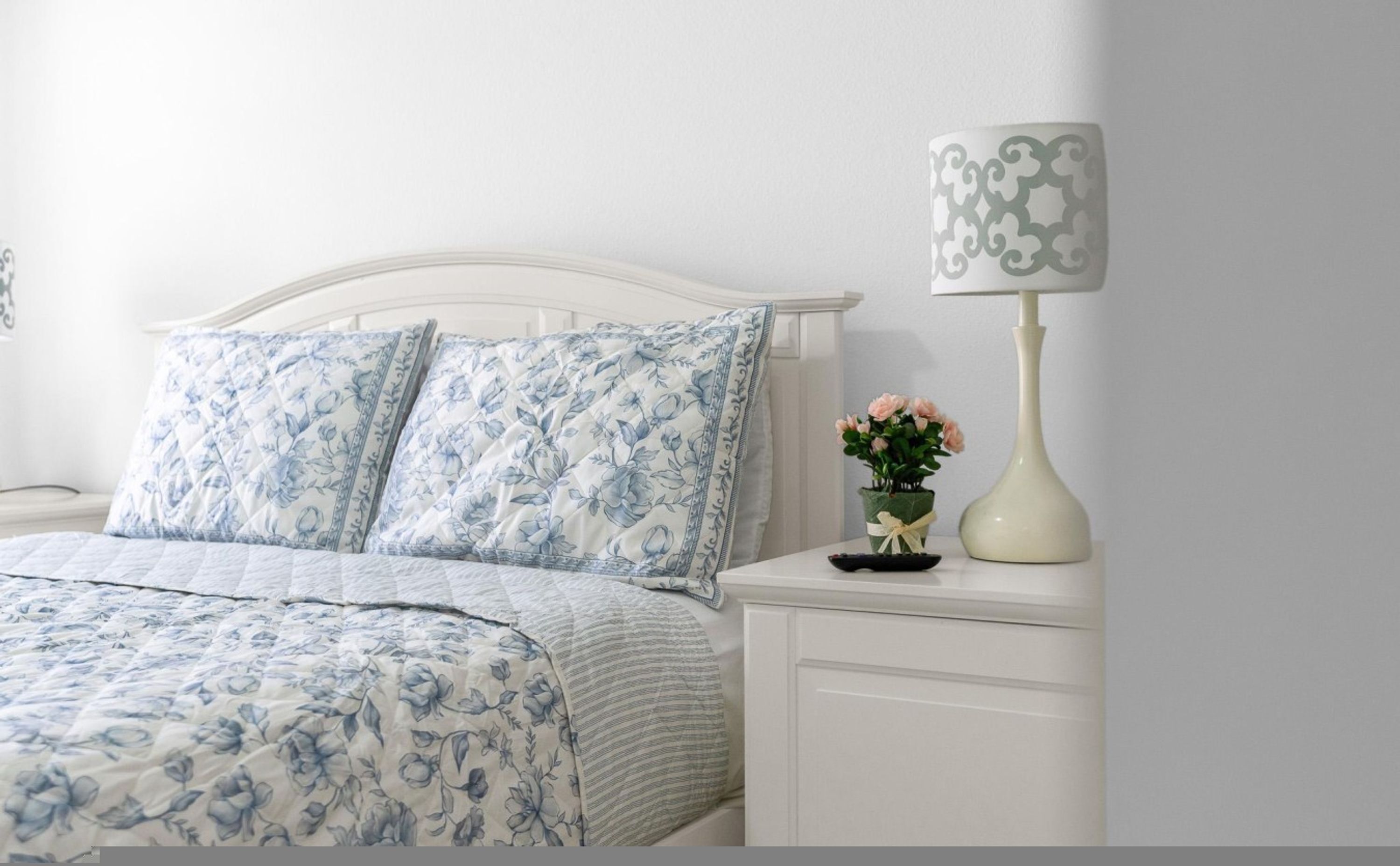 A bed with floral bedding, a bedside table with a lamp and a vase of flowers.