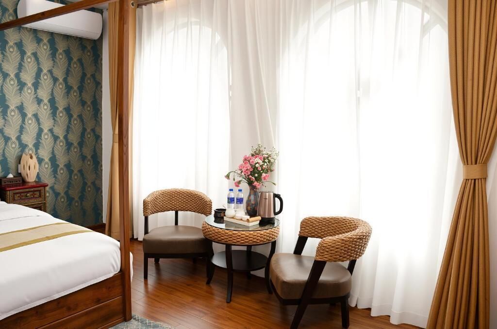Habitación de hotel con cama, dos sillones de mimbre, una mesita con un florero y una ventana con cortinas translúcidas.