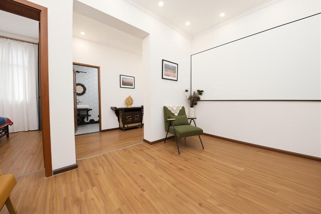 Una habitación con piso de madera, una silla verde, una mesita y un cuadro colgado en la pared, además de una puerta que da a otra habitación.
