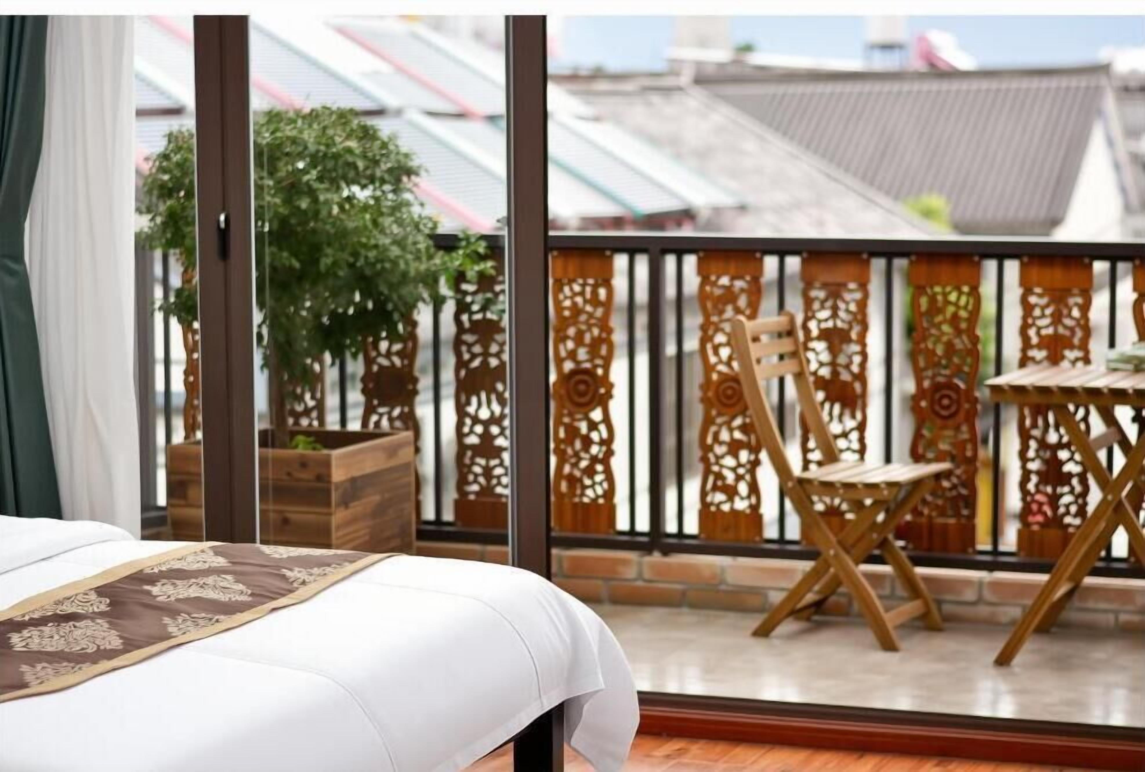 A room with a bed, a balcony with a wooden chair and table, and a view of buildings.